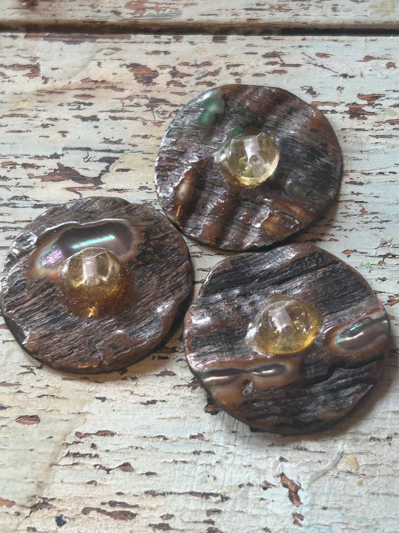 Op de afbeelding: Drie ronde, donkerbruine knopen met een getextureerde, houtachtige uitstraling. Elke knoop heeft een centrale, heldere, amberkleurige edelsteen. De knopen hebben iriserende highlights en zijn gerangschikt op een verweerd, lichtgekleurd houten oppervlak.