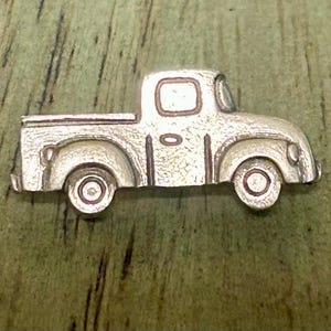 May include: A silver-toned metal pin shaped like a vintage pickup truck. The design features a cab, a truck bed, and two circular wheels with detailed rims. The surface of the metal has a textured, matte finish with defined outlines.