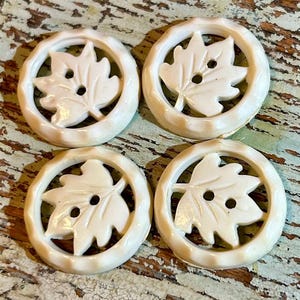 May include: Four off-white, circular buttons with a leaf design. Each button features a leaf shape in the center with two holes for sewing, surrounded by a textured ring. The buttons are arranged on a distressed, light blue and brown surface.