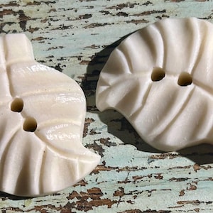 May include: Two off-white leaf-shaped buttons with two holes each. The buttons have a textured, veined design, and are set against a weathered, turquoise and brown wooden surface. These decorative buttons are ideal for sewing and craft projects.