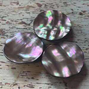 Op de afbeelding: Drie iriserende, ronde cabochons van parelmoer. De cabochons hebben een donkere basiskleur met glinsterende tinten roze, groen en paars. De cabochons zijn gerangschikt op een verweerd, lichtgekleurd houten oppervlak.