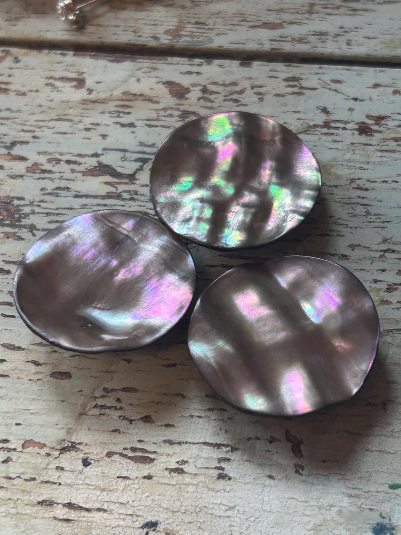 Op de afbeelding: Drie iriserende, ronde cabochons van parelmoer. De cabochons hebben een donkere basiskleur met glinsterende tinten roze, groen en paars. De cabochons zijn gerangschikt op een verweerd, lichtgekleurd houten oppervlak.
