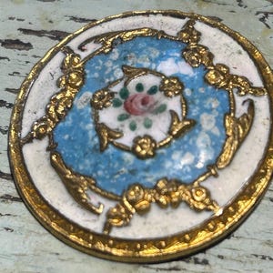 May include: A round, antique button with a gold-colored metal rim and ornate detailing. The center features a light blue background with a floral design and a pink rose. The outer edge is white.