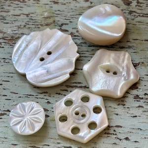 May include: A collection of iridescent, pearlescent buttons in various shapes. The buttons include a round, a flower-shaped, a scalloped, and a hexagonal design. The buttons have two or more holes for sewing and are made of a shiny, mother-of-pearl material.