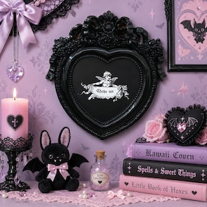 May include: A black heart-shaped frame with an ornate border containing a white graphic of a cherub holding a banner that reads "Choke me". The frame is displayed alongside a pink candle, a plush bat, a potion bottle, and three stacked books.