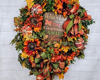 May include: A fall-themed wreath with a brown sign that reads "Harvest Thankful Fall Gratitude Family Leaves". The wreath is decorated with sunflowers, pumpkins, fall leaves, and a green and orange plaid ribbon.
