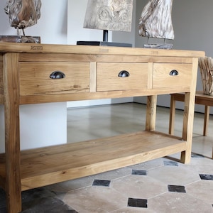 Puede incluir: Una consola de madera clara con tres cajones y un estante inferior. Los cajones tienen tiradores de metal oscuro. Esculturas decorativas de madera se encuentran encima. También se ven una pequeña silla de madera y una cesta tejida.