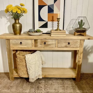 Może przedstawiać: Jasny drewniany stół konsolowy z trzema szufladami i dolną półką. Stół jest ozdobiony złotym wazonem z żółtymi kwiatami, oprawionym geometrycznym nadrukiem, świecznikiem i terrarium z kaktusem.