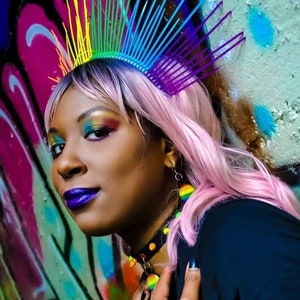 May include: A person with pink hair wearing a rainbow and purple crown, colorful eye makeup, and a black shirt with a rainbow choker.