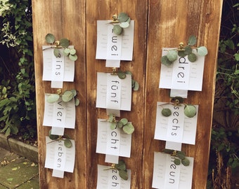 May include: A wooden sign with the words "find your seat" written in white paint. The sign is divided into three sections, each with a number written on a white card. The numbers are "eins", "zwei", "drei", "vier", "fünf", "sechs", "sieben", "acht", "neun", and "zehn".