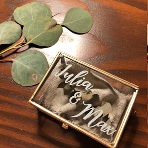 Puede incluir: Una caja de anillo de vidrio con un borde dorado y un forro de tela blanca. La caja tiene los nombres "Julia & Max" escritos en ella en caligrafía blanca.