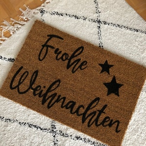 Könnte beinhalten: Eine braune Kokosmatte mit dem Text "Frohe Weihnachten" in schwarzer Schrift und zwei schwarzen Sternen.