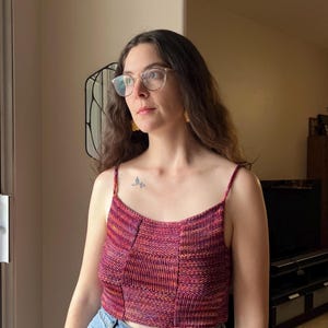 May include: A woman wearing a red, orange, and purple knit crop top with a scoop neck and thin straps. She is wearing blue jeans and has glasses on.