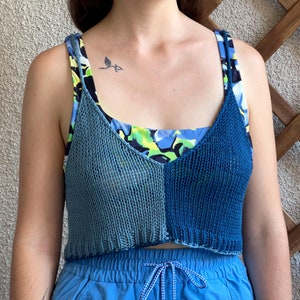 May include: A blue and teal knitted crop top with a contrasting floral print tank top underneath. The top has a relaxed fit and a slightly cropped length. The model is wearing the top with blue shorts.