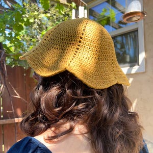 May include: A mustard yellow crochet bucket hat featuring a textured stitch pattern and a scalloped, wavy brim. The hat is constructed from yarn with visible vertical seams radiating from the crown down to the edge of the wide, flexible brim.