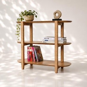 May include: A light brown wooden three-tier bookshelf with rounded oval shelves. The top shelf holds a potted green plant and a beige abstract sculpture. The middle shelf contains three books, and the bottom shelf holds a row of colourful books.