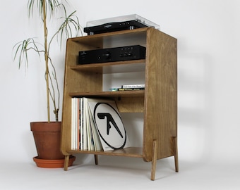 May include: A wooden record player stand with three shelves. The stand has a light brown finish and is made of plywood. The top shelf has a black record player on it. The middle shelf has a black amplifier on it. The bottom shelf has a stack of vinyl records and a book titled "Dark Side of the Moon" on it.