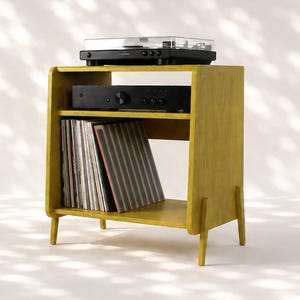 May include: A mustard yellow wooden media console with four tapered legs, featuring an open top shelf holding a black turntable, a middle shelf with a black audio amplifier, and a bottom shelf containing a collection of vinyl records.