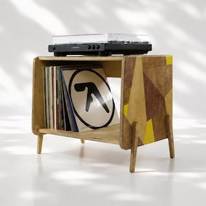 Puede incluir: Un soporte de madera para tocadiscos con cuatro patas, que presenta un patrón geométrico en marrón y amarillo en el panel lateral. El estante abierto contiene una colección de discos de vinilo, incluido uno con un logo en blanco y negro, coronado por un tocadiscos negro.