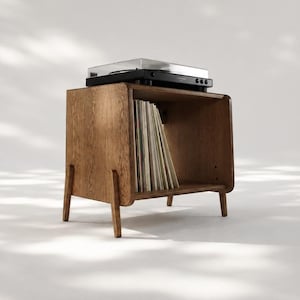 Puede incluir: Un soporte para tocadiscos de madera de color marrón medio con cuatro patas cónicas y un compartimento de almacenamiento abierto. El estante interior contiene una colección de discos de vinilo, con un tocadiscos negro en la parte superior.