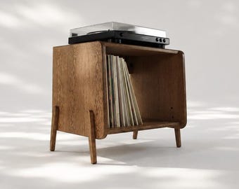 Puede incluir: Un soporte para tocadiscos de madera de color marrón medio con cuatro patas cónicas y un compartimento de almacenamiento abierto. El estante interior contiene una colección de discos de vinilo, con un tocadiscos negro en la parte superior.