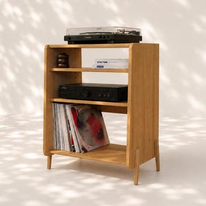 May include: A light brown wooden media console with four legs, featuring three open shelves. The top shelf holds a black turntable, the middle shelf contains a black audio amplifier and a white book, and the bottom shelf stores vinyl records.