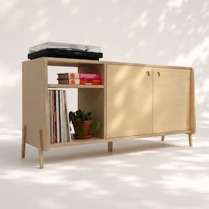 May include: A light-colored wooden media console with tapered legs, featuring an open shelf containing vinyl records and a small potted plant, a second shelf with two books, and a closed cabinet section with two circular cut-out door handles.