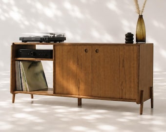 May include: A rectangular wooden media console featuring a natural wood grain finish, supported by four tapered legs. The unit includes an open shelf section for a turntable and vinyl records, and a closed cabinet section with two circular handles.