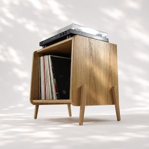 Puede incluir: Un soporte para tocadiscos de madera de roble claro con cuatro patas cónicas y un compartimento de almacenamiento abierto. Un tocadiscos negro se sitúa en la superficie superior, mientras que una colección de vinilos se organiza verticalmente.