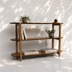 May include: A three-tier wooden bookshelf with rounded ends and a medium brown finish. The shelves hold two small potted green plants, a small dark wooden object, and four books with white covers, including titles like Kinfolk and Brutus.