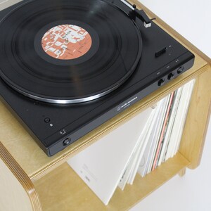 May include: A black record player with a record on the turntable sits on a light wood shelf with a stack of records underneath. The record player has a silver tone arm and a clear dust cover.