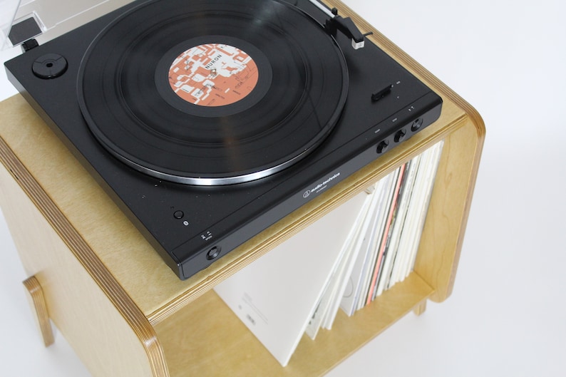 May include: A black record player with a record on the turntable sits on a light wood shelf with a stack of records underneath. The record player has a silver tone arm and a clear dust cover.