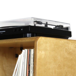 May include: A black turntable with a clear dust cover sits on top of a light wood record player stand. The stand has a shelf below the turntable that is filled with vinyl records.