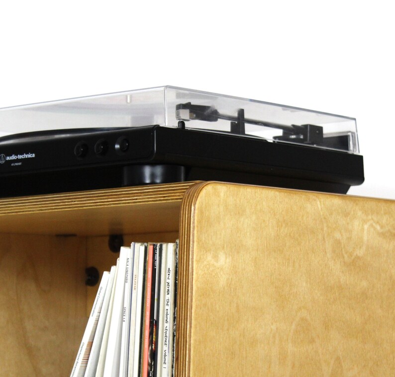May include: A black turntable with a clear dust cover sits on top of a light wood record player stand. The stand has a shelf below the turntable that is filled with vinyl records.