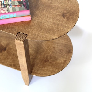 May include: A two-tiered wooden side table with a rounded top and bottom shelf. The table is made of light brown wood and has a natural wood finish. The top shelf has a stack of three books on it, with the top book titled "FRESH".