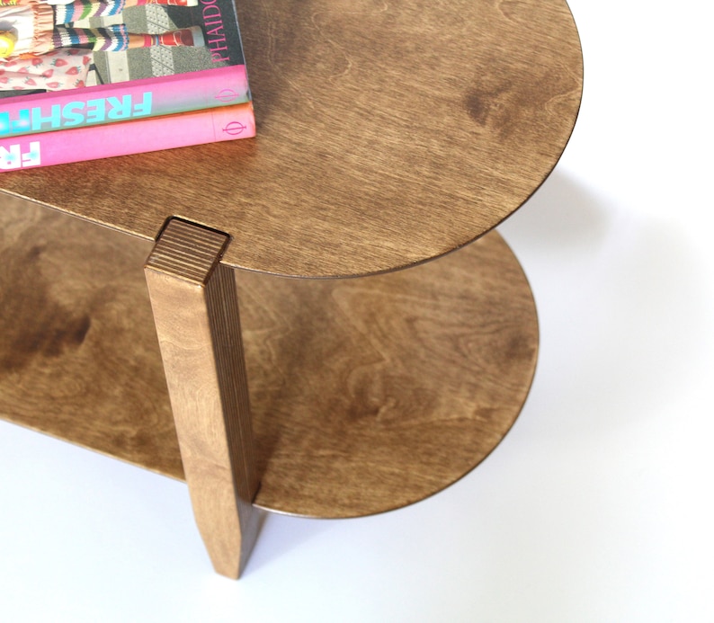 May include: A two-tiered wooden side table with a rounded top and bottom shelf. The table is made of light brown wood and has a natural wood finish. The top shelf has a stack of three books on it, with the top book titled "FRESH".
