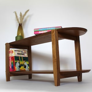 May include: A wooden end table with a rounded top shelf and a lower shelf. The table has a natural wood finish and is decorated with a vase of dried flowers and three books. The books are titled "Fresh", "Now!" and "Japanese Graphics".