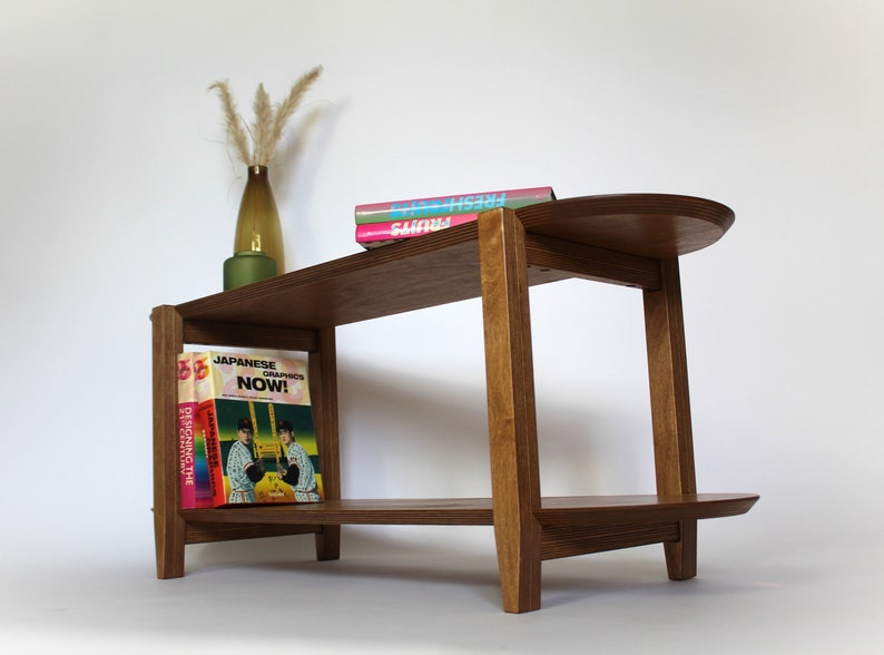 May include: A wooden end table with a rounded top shelf and a lower shelf. The table has a natural wood finish and is decorated with a vase of dried flowers and three books. The books are titled "Fresh", "Now!" and "Japanese Graphics".