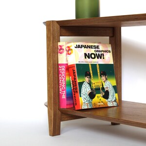May include: A wooden bookshelf with three shelves. The top shelf has a stack of books. The middle shelf has three books, one of which is titled "Japanese Graphics Now!" The bottom shelf is empty.