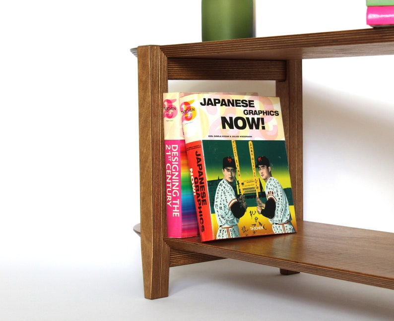 May include: A wooden bookshelf with three shelves. The top shelf has a stack of books. The middle shelf has three books, one of which is titled "Japanese Graphics Now!" The bottom shelf is empty.