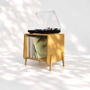 May include: A light-toned wooden record player stand with four tapered legs and an open storage compartment filled with vinyl records. A black turntable with a clear plastic dust cover is positioned on the top surface of the wooden stand.