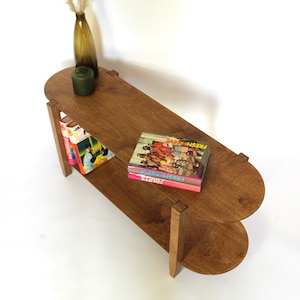 May include: A two-tiered wooden side table with rounded edges. The table has a dark brown stain and features two shelves. The top shelf has a green vase and a stack of books with the title "FRESH" on the top book.