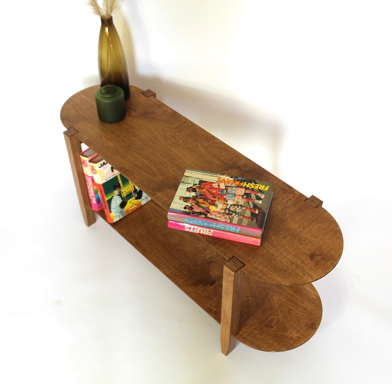 May include: A two-tiered wooden side table with rounded edges. The table has a dark brown stain and features two shelves. The top shelf has a green vase and a stack of books with the title "FRESH" on the top book.
