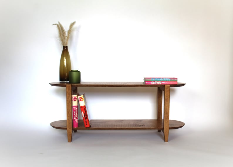 May include: A two-tiered wooden coffee table with a light brown finish. The table has oval-shaped shelves and a simple, modern design. It is decorated with a vase of dried grass and a small green vase.