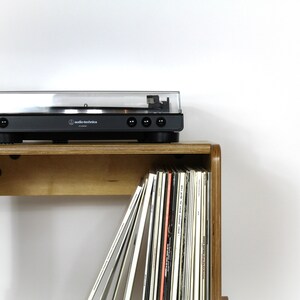 May include: A wooden record shelf with a turntable on top. The shelf has a built-in compartment for storing vinyl records. The turntable is black with a clear lid and the text "audio-technica" printed on the front.