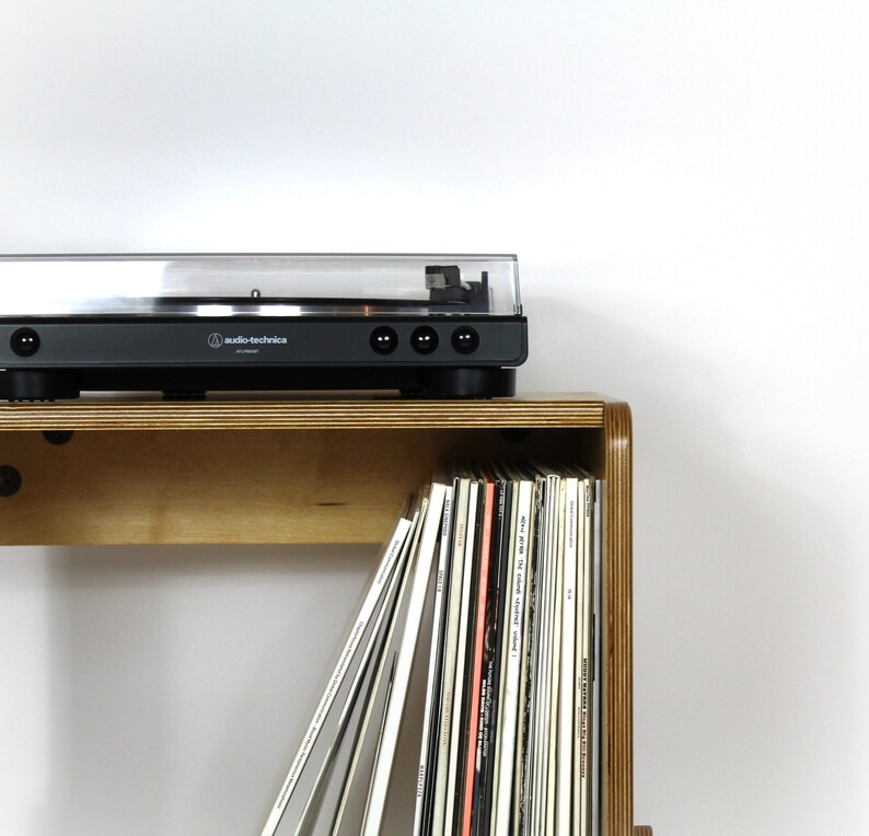 May include: A wooden record shelf with a turntable on top. The shelf has a built-in compartment for storing vinyl records. The turntable is black with a clear lid and the text "audio-technica" printed on the front.
