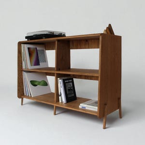 May include: A medium-toned wooden storage unit with rounded corners and four tapered legs. The unit features two horizontal shelves divided into four sections, currently holding vinyl records and books, with a record player on the top surface.
