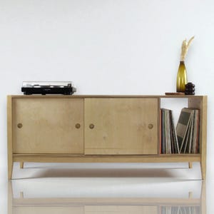 May include: A light wood media unit with sliding doors and a shelf for vinyl records. A black turntable sits on top, with a decorative vase and objects. The unit has a mid-century modern design, with a height of approximately 60 cm.