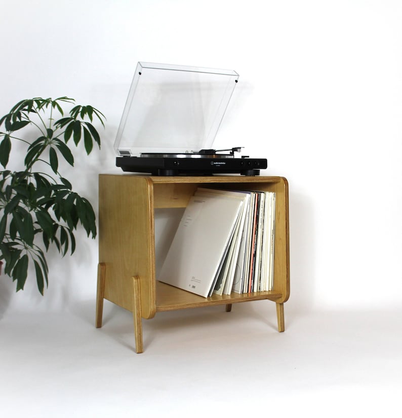 May include: A light wood record player stand with a turntable and a shelf for vinyl records. The stand has a clear plastic cover for the turntable and four legs.