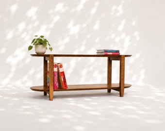 May include: A brown wooden console table with two horizontal shelves and four legs. A small green potted plant sits on the left side of the top shelf. Two books are stacked on the right side of the top shelf, and two books stand on the bottom shelf.